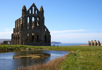 This landscape photograph captures Whitby Abbey, a historic church ruin located in Whitby, Yorkshire, as seen from the south east. The abbey stands prominently against a bright blue sky, reflecting in a small pond in the foreground. Taken in the early afternoon during spring, the image highlights the dramatic Gothic architecture of Whitby Abbey, with the North Sea and the coastal cliffs of Yorkshire visible in the background. The clarity of the scene and the fresh greenery suggest a mild and pleasant spring day, showcasing both the cultural heritage of Whitby and the scenic beauty of Yorkshire.